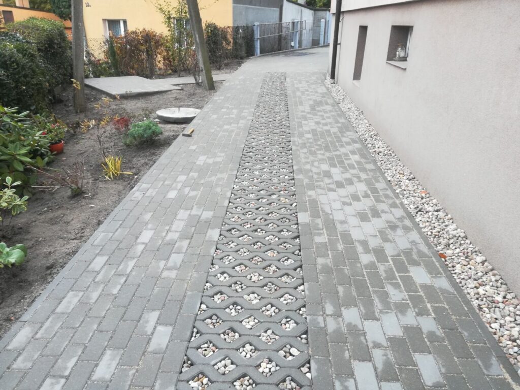 Władysławowo - Układanie kostki brukowej w ogrodzie, z widoczną ścieżką wykonaną z szarej kostki brukowej oraz dekoracyjnym wzorem z otworami na roślinność. Prace brukarstwa obejmują estetyczne i funkcjonalne elementy krajobrazu.