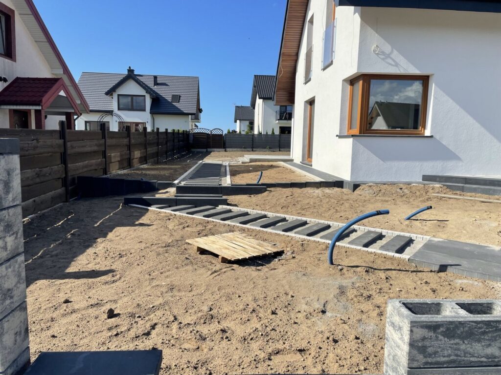Kosakowo - Układanie kostki brukowej w nowo budowanym ogrodzie, z widocznymi przygotowaniami do brukarstwa. Zarysowane ścieżki z kostki brukowej i elementy infrastruktury w tle.