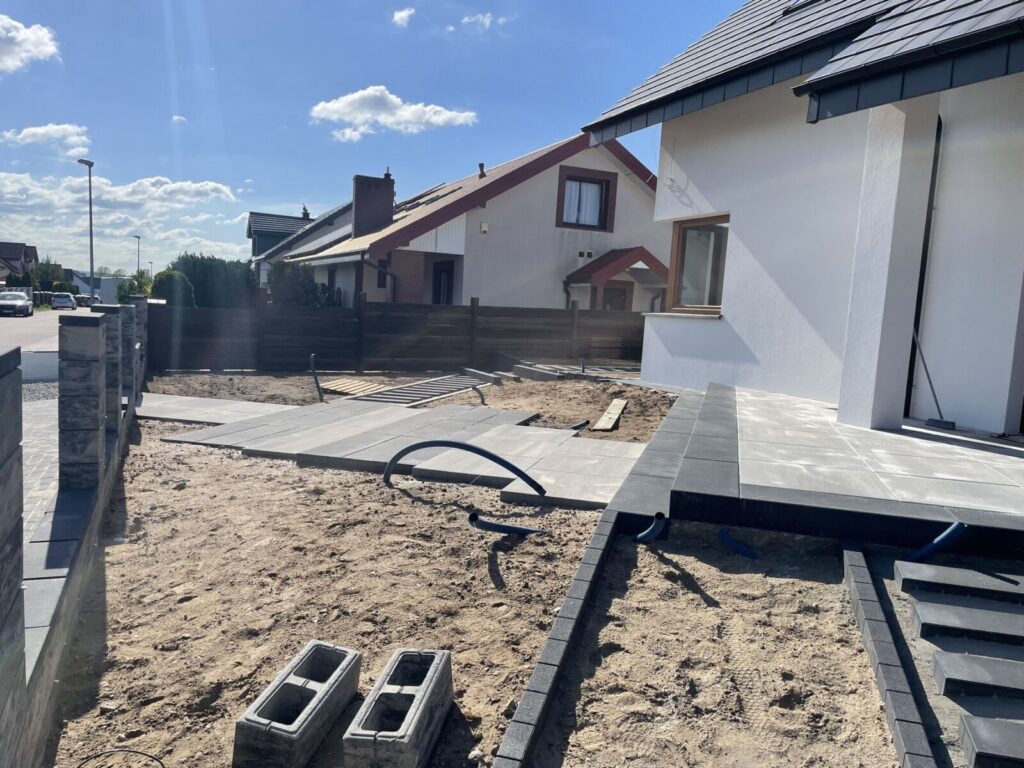 Hel - Widok na plac budowy z układaniem kostki brukowej, gdzie widać świeżo ułożone płyty oraz przygotowane miejsce na dalsze brukarstwo. W tle znajdują się domy jednorodzinne, a teren jest w trakcie zagospodarowania.