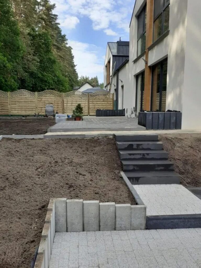 Widok na nowo ułożoną kostkę brukową w ogrodzie, z wyraźnie zaznaczonymi ścieżkami i schodami. Układanie kostki brukowej zapewnia estetyczny wygląd przestrzeni zewnętrznej, a brukarskie prace są w pełni widoczne w tle.