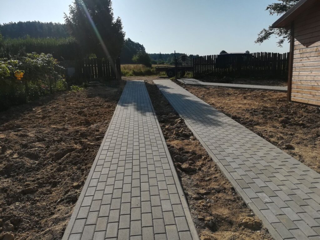 Kosakowo - Ścieżki z kostki brukowej w ogrodzie, pokazujące układanie kostki brukowej na świeżo przygotowanej ziemi. W tle widoczne rośliny i drewniany domek. Idealne dla osób interesujących się brukarstwem i aranżacją przestrzeni zewnętrznej.