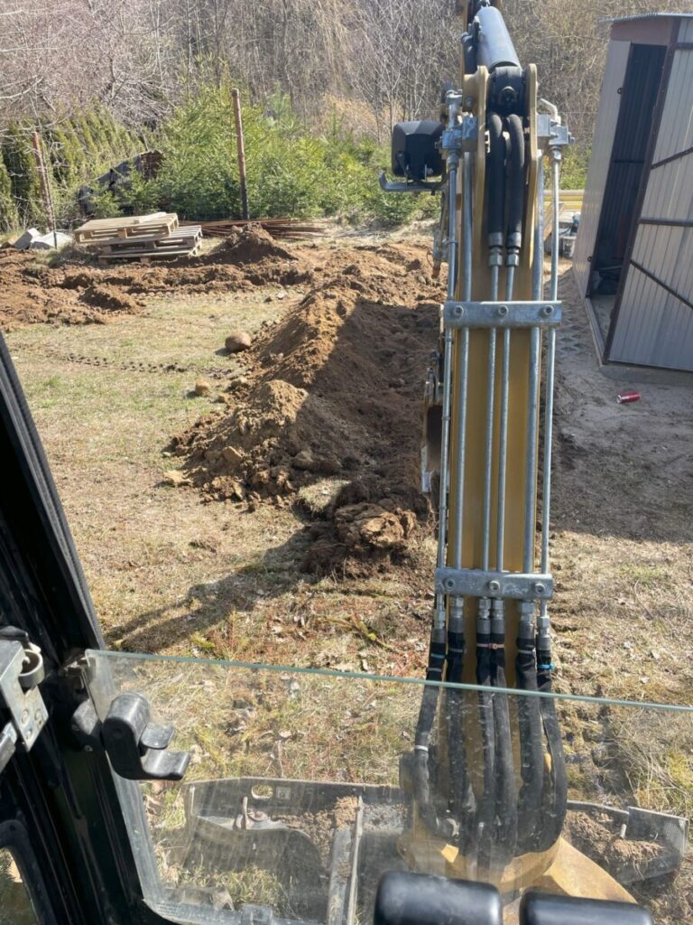 Widok z kabiny koparki na plac budowy, gdzie przygotowywana jest ziemia pod układanie kostki brukowej. W tle widać wykopany teren oraz materiały budowlane, co wskazuje na prace związane z brukarstwem. - Kosakowo