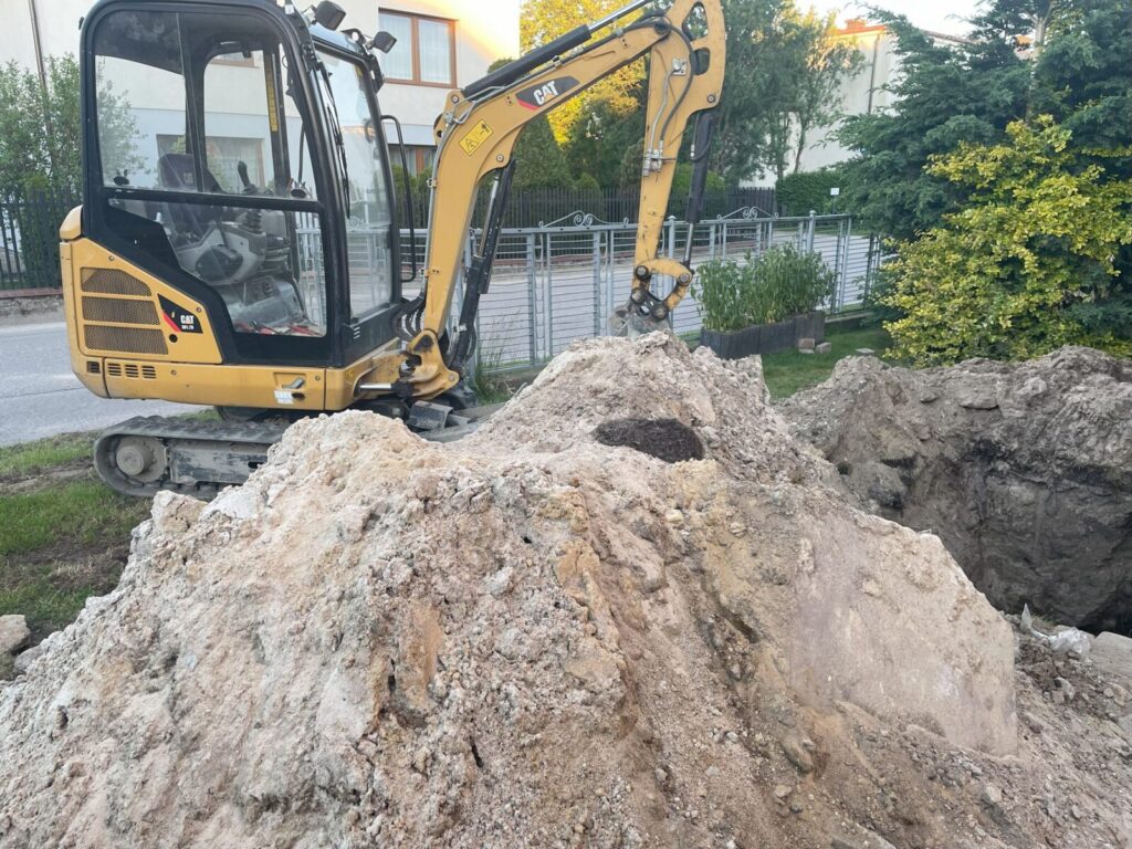 Hel - Maszyna budowlana przygotowująca teren pod układanie kostki brukowej. W tle widoczny wykop oraz hałda ziemi, co sugeruje prace związane z brukarstwem.
