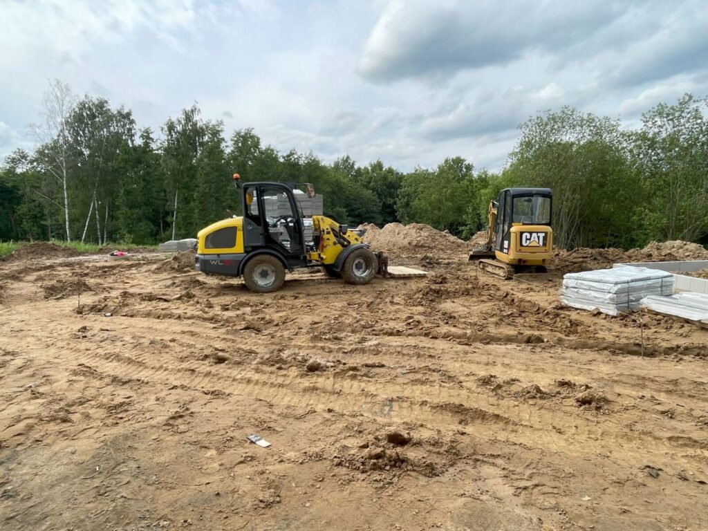 Gniewino - Prace budowlane na terenie przygotowanym do układania kostki brukowej, z użyciem maszyn budowlanych do brukarstwa. W tle widoczne materiały do kostki brukowej oraz zalesienie.