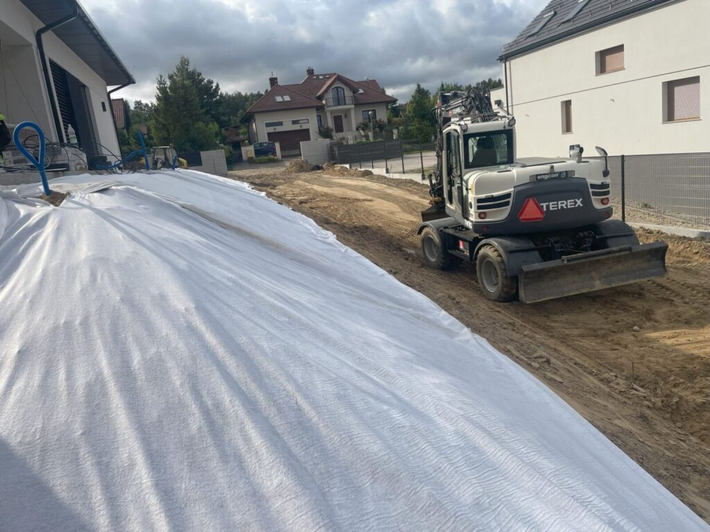 Kosakowo - Maszyna budowlana Terex przygotowuje teren do układania kostki brukowej, z widoczną warstwą geowłókniny w tle, co wskazuje na prace brukarstwa w nowym osiedlu.