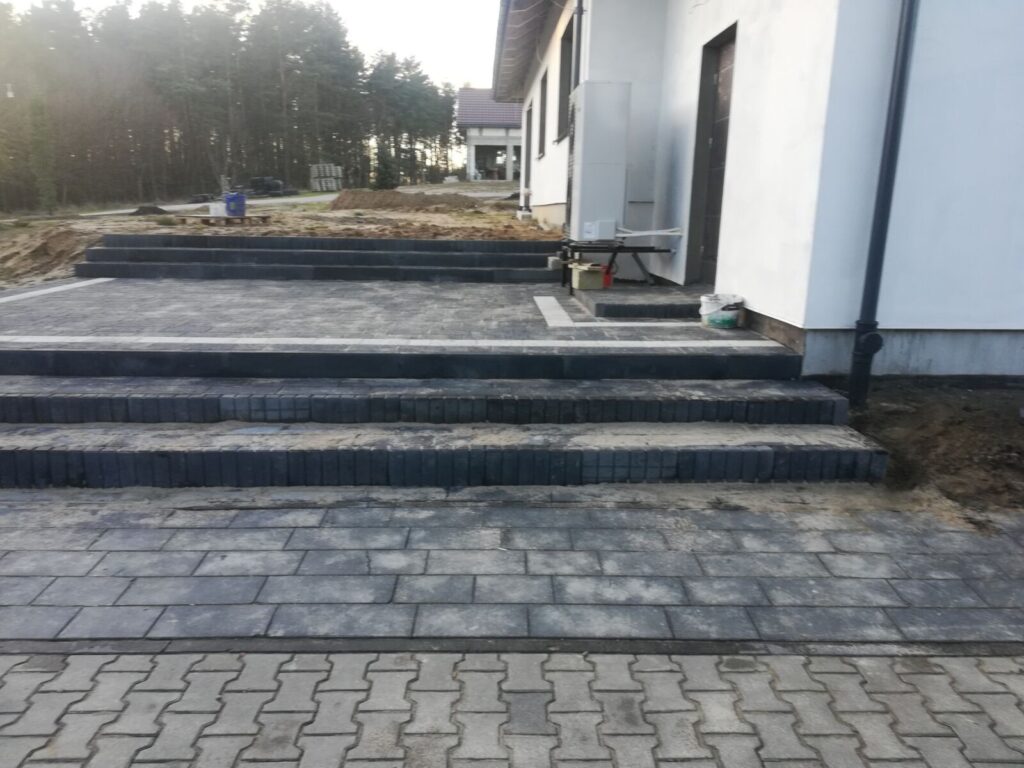 Gniewino - Układanie kostki brukowej na schodach i chodniku wokół nowo wybudowanego domu. Widoczna kostka brukowa o ciemnym kolorze, tworząca estetyczne i funkcjonalne przestrzenie. Prace brukarstwa w tle, z widocznymi przygotowaniami do dalszych etapów budowy.
